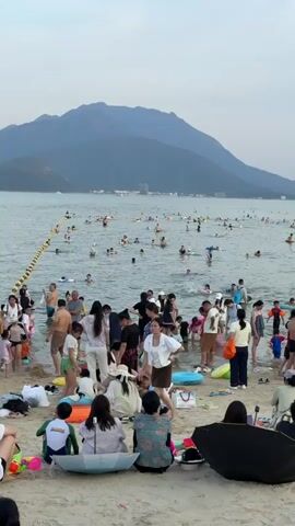 The video depicts a crowded beach with many people swimming and playing in the water. The water is calm and shallow near the shore, with a yellow rope marking a boundary. Numerous individuals, including children and adults, are seen wading, swimming, and floating on inflatable devices. On the sandy shore, people are sitting, standing, and interacting. There are various objects scattered on the sand, such as beach towels, bags, umbrellas, inflatable rings, and toys. The background features a calm sea extending towards a hazy horizon, with mountains visible in the distance under a cloudy sky. The overall impression is a bustling recreational scene on a summer day.
