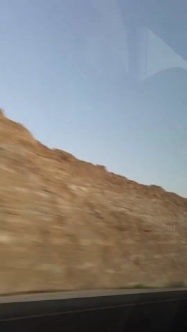 A moving vehicle's perspective captures a blurry, tan-colored hillside against a light blue sky. The hillside occupies the majority of the frame, with a dark horizontal line, likely a road or barrier, at the bottom. The sky appears clear with faint reflections on the vehicle's window. The overall impression is of driving through a barren, desert-like landscape.