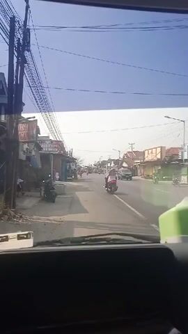 A daytime view from inside a vehicle looking forward through the windshield. The road ahead is paved and appears to be a two-lane street with traffic. On the left side of the road are buildings, some with storefronts, and utility poles with numerous electrical wires. A few motorcycles are parked on the left side. On the right side of the road, there are more buildings and some vegetation. Several vehicles, including cars and motorcycles, are visible on the road, moving in both directions. The sky is clear and blue.