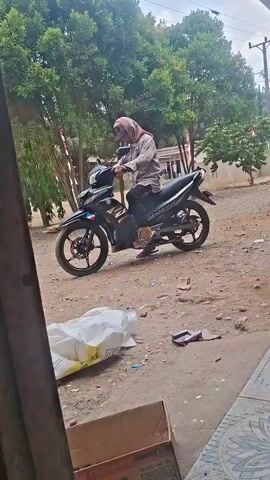 A person wearing a hijab and a light-colored long-sleeved shirt is sitting on a black motorcycle. The motorcycle has a blue and white emblem on its front. The person is holding the handlebars and looking forward. The motorcycle is parked on a dirt road. In the foreground, there is a large white bag and a cardboard box. In the background, there are trees and some buildings. The scene appears to be outdoors in a rural or semi-rural setting.