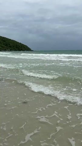 A tranquil beach scene unfolds under a cloudy sky. Gentle waves lap at the sandy shore, creating foamy patterns as they recede. In the distance, a small, tree-covered island sits amidst the turquoise water. The overall atmosphere is serene and peaceful, inviting a sense of calm.