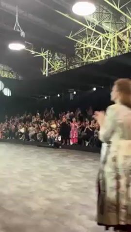 A group of women models, all smiling and wearing different colorful outfits, walks down a fashion runway in front of a crowd. The event appears to be indoors in a large open space with a stage and lighting. A screen displays images of women in the background. The models clap and interact with each other as they proceed down the runway. Electronic dance music plays in the background.