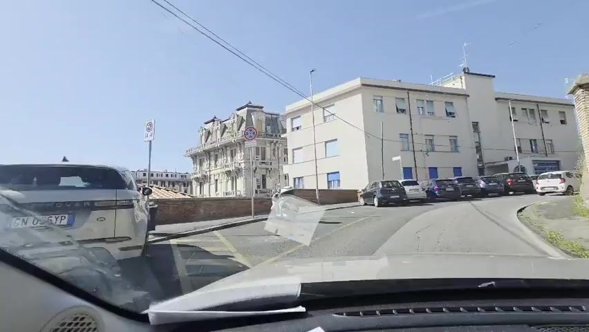 A POV shot from a vehicle shows a street with parked cars and buildings. The vehicle is moving slowly down a road. On the left is a cream-colored building with a blue trim around the windows and what appears to be a garage door. To the left of that building is a smaller, older building with a decorative mansard roof and ornate architectural details. Further down the street, on the right, is a steep, rocky embankment covered in vegetation. The road is lined with parked cars, and there is a white car in the distance. The sky is clear and blue.