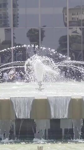The video displays a close-up shot of a multi-tiered fountain. Water jets upward from the center, creating a misty spray. Smaller streams of water cascade down the sides of the tiered structure. In the background, a modern building with large, reflective glass windows is visible, with reflections of trees and urban structures. Blurry figures of people can be seen in the background, some gathered near the fountain. The lighting suggests it is daytime.