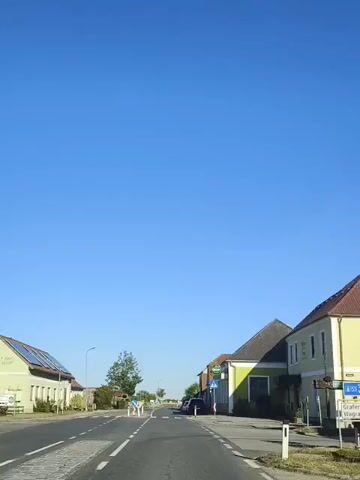 The video shows a street scene on a clear day with a bright blue sky. The camera is mounted on a vehicle moving forward on a paved road. On the left side of the road, there are buildings, one with solar panels on its roof, and a few trees. On the right side of the road, there are buildings, one painted lime green with a sign that appears to say "Gösser." Several cars are parked along the right side of the road, including a red SUV and a white sedan. There is a crosswalk marked on the road in front of the lime green building. Traffic signs and bollards are visible along the road. The overall environment appears to be a village or small town.
