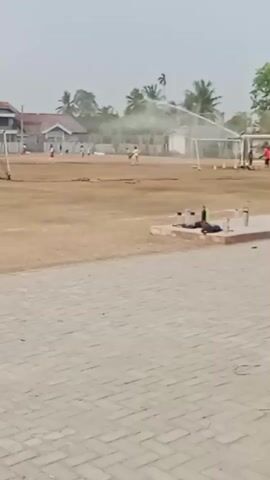 This video shows a dusty, outdoor sports field with several soccer goals. In the distance, a group of young children are playing soccer. A few adults are also present, possibly supervising or participating. The ground appears to be dry and dusty, with some patchy grass. The sky is overcast. There are some houses and trees visible in the background. The video is filmed from a slightly elevated perspective, looking down onto the field.