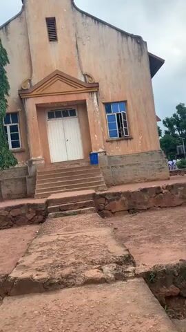 The video shows the exterior of a peach-colored church with white double doors at the entrance, accessible by a set of stairs. To the left of the entrance are two windows, and to the right is one window, all with blue panes. Above the entrance, a triangular pediment is visible. The church has a sign above the pediment that reads "1948." The building is situated on a reddish-brown dirt ground, with some sparse vegetation. A tree is visible to the far left. The sky is overcast. There is a blue plastic bin next to the stairs.