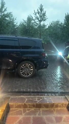 A dark blue SUV is parked on a wet brick driveway. A person in a bright yellow raincoat is standing near the open back of a dark vehicle, possibly a golf cart or small utility vehicle, to the right of the SUV. The person appears to be interacting with the vehicle. It is raining, and the ground is wet, with water pooling and running in gutters. Trees are visible in the background, and string lights are hanging overhead. There are also small outdoor lights on the ground. The sky is overcast. The scene appears to be an outdoor residential or recreational area.