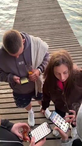 A group of three people are standing on a wooden dock or pier over a body of water. Two of them are holding cell phones, with one of them attempting to airdrop something to the other. The person receiving the airdrop raises their hand up with their phone as if to make the connection stronger. The people are young adults wearing casual clothing. One is holding a glass of orange liquid. The dock has horizontal wooden planks, and there are light ripples on the water.
