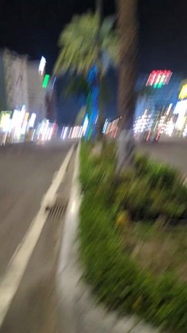 The video shows a street in a city at night. Palm trees line the street, decorated with colored lights. Buildings are visible on both sides of the street, illuminated with various lights. Cars and motorbikes are driving on the road. The camera is positioned close to the ground, giving a low-angle view of the scene.