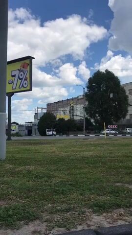 The video captures a daytime urban street scene in San Martín de Porres, Peru, panning from left to right. The sky is partly cloudy.

Initially, a large, somewhat dilapidated building is visible, featuring a sign with the word "ELEPHANT" on it, along with a yellow star graphic. A white van is parked nearby. To the left, part of a large billboard is cut off from view. A grass median separates the viewer's perspective from the multi-lane road.

As the camera pans right, a large, dense green tree briefly obstructs part of the view. Beyond the tree, cars of various colors (silver, red, white) are seen driving on the road. A red and white chevron traffic sign is visible near the median.

Further panning reveals a two-story commercial building with multiple shopfronts on the ground floor. The upper facade of this building appears weathered and partially stripped. Some visible text on the shop windows includes "MK" and possibly other words, which appear to be in Cyrillic script. People are seen walking on the sidewalk in front of these shops.

The pan concludes with a view of a much taller, dark-colored multi-story building with numerous windows, standing behind and to the right of the two-story structure, typical of an older city high-rise. Overhead wires and street lamp poles are present throughout the scene. The overall impression is one of a busy commercial area.