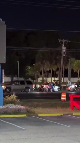 At night, a group of motorcycles are stopped at a traffic light, behind a silver SUV and ahead of a white sedan. The scene is lit by streetlights. Palm trees line the road. A large red chair sits in the foreground, and a sign for a gas station is visible on the right. The camera pans to the right as the motorcycles start to move.