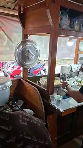 The video shows a person preparing food at a street food cart. The cart is made of wood and has a metal roof. There are stacks of bowls and plates on shelves. The person is wearing a black shirt and a peci cap. They are using a ladle to serve food from a pot. There are also some plastic cups and a bottle of water on the counter. In the background, there are motorcycles parked. The environment appears to be outdoors, possibly a street or market.