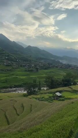The video shows a sweeping panoramic view of a lush green valley filled with terraced rice paddies. The sun is setting, casting a warm glow over the landscape and highlighting the rolling hills and mountains in the background. A small village is nestled in the valley, with houses scattered among the rice fields. A river flows through the valley, adding to the serene beauty of the scene. In the foreground, the rice paddies are more pronounced, with distinct steps and curved patterns. The sky is filled with dramatic clouds, painted with hues of orange and pink from the setting sun. The overall atmosphere is one of tranquility and natural beauty.