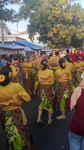 A group of dancers in traditional Indonesian costumes are performing outdoors. The dancers wear bright yellow tops with intricate gold embroidery and dark patterned skirts over bright green pants. They have their hair styled with flowers. The performance is taking place on a paved street, with onlookers standing on the sides. In the background, there are market stalls with blue awnings, trees, and buildings. The lighting suggests it is daytime, possibly late afternoon. The dancers appear to be in motion, engaged in a choreographed routine. One dancer in the foreground has their back to the camera, while others are facing partially or fully away. A white motorcycle is parked on the left side of the street.
