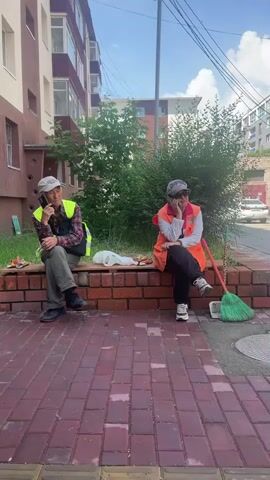 Two individuals are sitting on a low brick wall. Both are wearing hats and vests, suggesting they may be workers. The person on the left is wearing a reflective yellow vest over a plaid shirt and dark pants, with dark shoes. They are holding a phone to their ear and appear to be speaking. In front of them are some food items and a white plastic bag. The person on the right is wearing an orange vest over a white shirt and dark pants, with white sneakers. They are also holding a hand to their face, possibly resting their chin. A green broom is leaning against the brick wall to their right. The background shows a building, trees, and a street with a car parked in the distance. The ground in front of the wall is paved with red bricks, and there are yellow tactile paving tiles at the edge of the walkway. The scene is outdoors during daylight.