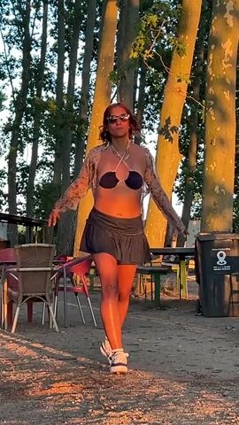 A person with reddish-brown hair, wearing sunglasses, a dark blue bikini top, a sheer floral long-sleeved cardigan, and a dark, pleated mini-skirt, is dancing outdoors. The person is also wearing white sneakers with grey accents and white socks. The ground is sandy and covered with fallen leaves. In the background, there are several tall trees with sunlight filtering through their leaves, casting long shadows. There are also picnic tables, chairs, and trash cans visible. The overall setting appears to be a park or a recreational area.