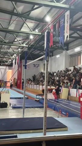 A young female gymnast in a red leotard stands on a balance beam, her arms raised in celebration, in a gymnasium. Spectators fill the stands behind her. A Turkish flag hangs on the wall to her left. Two horizontal bars are visible in the foreground. The ceiling is high and industrial.