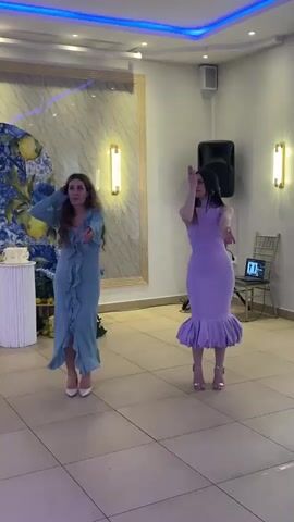 Two women are dancing indoors in a formal setting. The woman on the left has long brown hair, wearing a light blue dress with ruffles down the front and white high heels. The woman on the right has long dark hair and is wearing a form-fitting purple dress with a ruffled hem and gold high heels. They both hold their hands to their heads and then down to their waists in a synchronized dance. In the background is a wedding cake on a stand, a tall framed painting, a speaker, a laptop on a chair, and modern sconces. Ceiling lights cycle through different colors. The room is brightly lit. The floor is tiled.