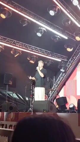 The video shows a man on a stage holding a microphone and a phone. He is wearing a black shirt and white pants. There are lights and speakers on the stage. The man appears to be counting down from ten. The background is dark with some colorful graphics. There are people in the foreground, out of focus.