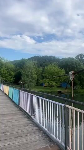 The video captures a static view from a wooden bridge, looking out over a body of water. The bridge features a railing that transitions from light blue and yellow to white. Thick metal cables, part of the bridge's structure, are visible stretching across the frame. On the right side of the bridge, a unique metal object resembling a mask or face is attached to one of these cables.

Beyond the water, the scene is dominated by lush green trees and vegetation, indicating a vibrant natural environment. On the far bank, several small, brightly colored structures, notably blue and yellow, are nestled among the trees. In the distance, high on a tree-covered hill, a building with a distinctive conical roof, possibly a castle or watchtower, can be seen peering through the foliage. The sky is partly cloudy, suggesting a bright daytime setting. The overall atmosphere is serene and picturesque. This scene is located in Kamianytsia, Ukraine.