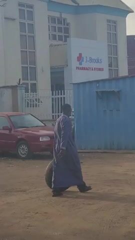 A man in a long blue garment walks from left to right across a dirt road, carrying a tire around his waist. Behind him is a building with a sign that reads "J-Brooks Pharmacy & Stores." To the right of the man is a small blue metal structure and a gold car. The sky is overcast.