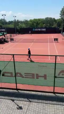 Two people are playing tennis on a red clay court on a sunny day. The camera angle is from above, slightly behind the back fence of the court. One person is wearing blue and serving. The court is surrounded by a green fence with the word HEAD prominently displayed in white lettering. There are buildings and trees in the background. The words UTR PRO TENNIS are visible on a screen in the background.
