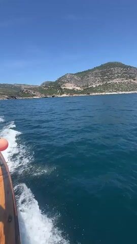A boat moves through clear blue water on a sunny day, leaving a white wake behind it. The camera is positioned at the back of the boat, looking towards the water and the distant coastline. To the left, a wooden part of the boat and a small orange buoy are visible. In the background, a large, rocky island or mainland with sparse green vegetation rises from the sea under a bright, cloudless blue sky. The water exhibits varying shades of blue and turquoise, indicating depth. The scene suggests a pleasant day out on the water in a coastal area, likely near Menderes, Turkey.