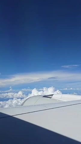 This is a first-person view from an airplane window. The camera is pointed towards the right wing of the plane and out into the sky. The sky is a deep blue with white, fluffy clouds spread out below the plane. The wing is white and dominates the foreground, casting a shadow that extends into the lower left corner of the frame. A portion of the airplane engine nacelle is visible on the right side, nestled against the wing. The camera appears to be stationary, and there are no visible people or distinct actions occurring. The environment is the upper atmosphere.