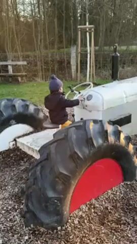 A young child, facing away from the camera, sits on a gray tractor, which appears to be a playground structure. The child wears a dark jacket, yellow pants, and a dark blue knit cap. The child is holding onto the tractor’s steering wheel, as if driving. The tractor has large black tires with a red painted section on the sides. The ground is covered in wood chips. A wooden bench and a small tree are visible in the background, along with other trees in the distance.