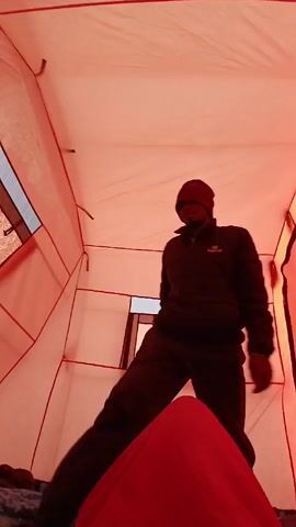 Inside a tent with red-tinted light, a person wearing a dark jacket, pants, and a dark beanie stands and looks down. The tent has several small, rectangular windows. The person picks up a blue and gray camouflage-patterned blanket. The tent poles are visible. There is a red sleeping bag at the bottom of the frame. The person seems to be tidying up or packing items in the tent.
