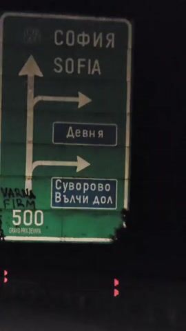 The video opens with a close-up of a green road sign with white text and symbols. Some of the text is in English, including "VARNA" and "500". There are also some handwritten markings in black on the sign. The camera then pans away to reveal a dark, nighttime scene of a highway. Two cars with their taillights illuminated are driving ahead on the highway. The video continues with a view of the road ahead, with white lines marking the lanes. The scene is dark, and the only significant light sources are the taillights of the cars and some distant lights. The video appears to be shot from inside a moving vehicle.