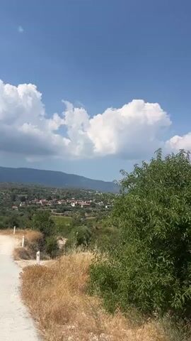 The video shows a rural landscape on a sunny day with a clear blue sky and fluffy white clouds. A paved road curves into the foreground from the left, transitioning to a dirt path that leads towards a village nestled in the valley below. The village consists of small buildings with red-tiled roofs, surrounded by lush green trees and olive groves. Rolling hills and mountains form the backdrop, with more scattered vegetation on their slopes. In the foreground, dry grass and brush line the sides of the path. There are no people or animals visible in the video.