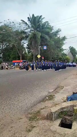 A marching band, composed of approximately 30 individuals wearing blue uniforms with white sashes, walks in formation on a paved road. The band members appear to be carrying brass instruments. They are moving from left to right in the frame. To the left of the road, several motorcycles and a few individuals are visible, as well as a red and blue tent. Further in the background, there are trees, including a prominent palm tree, and utility poles. The sky is overcast. The foreground shows a rough, concrete structure with a drain opening. The overall scene suggests a parade or public event taking place outdoors.
