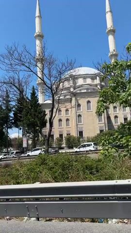 A camera pans across a city street on a clear, sunny day. In the foreground, a metal barrier separates the road from a grassy area. A highway is visible, with cars driving by. To the left, a mosque with white minarets stands tall. Further on, a cylindrical building with a domed roof and vertical windows is visible amidst lush greenery. The camera continues to pan, revealing more trees and the blue sky.