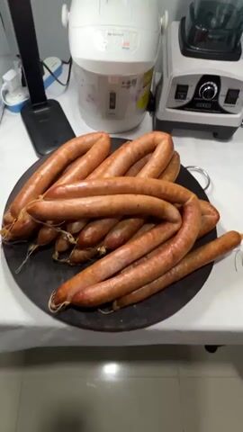 This is a close-up video of a pile of sausages. The sausages are linked together with twine and are a reddish-brown color. They are arranged on a dark circular platter, which is placed on a white surface. In the background, there is a white rice cooker, a black desk lamp, and a white blender. The lighting appears to be artificial and from above. The overall scene suggests food preparation or storage.
