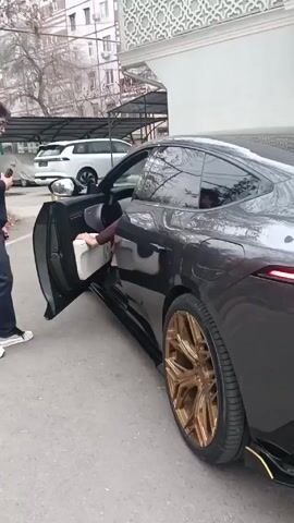 Here is a detailed description of the video:

The video shows two men standing next to a black car with gold-colored wheels parked on an asphalt surface. The man closer to the camera is wearing a navy blue hooded jacket, dark pants, and white and black sneakers. He has dark hair and glasses and is holding what appears to be a small item in his hand. The other man is exiting the car. He is wearing a maroon sweater, brown pants, and black shoes. The car door is open.

In the background, there is a parking area with a covered structure and several parked vehicles, including a white SUV. A building with white walls and a patterned design is visible in the background. Bare trees are also visible in the background, suggesting it might be late fall or winter.

The camera follows the man in the blue jacket as he turns to look at the man exiting the car, and then back to the car. The man exiting the car adjusts himself and the open door. The audio contains indistinguishable speech.