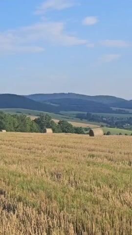 The video shows a rural landscape on a clear day. The foreground is a harvested field with stubble, and in the midground, there are two round hay bales. Beyond the field, there are rolling green hills with trees and a village nestled in the valley. In the distance, more hills fade into a hazy blue under a sky with scattered white clouds. The camera pans slowly to the right.