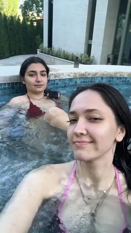 Two young women are in a hot tub, presumably outdoors given the green foliage and building in the background. The woman in the foreground, wearing a pink bikini top and necklaces, smiles and looks at the camera, her hair wet. The woman in the background, wearing a burgundy bikini top, is partially submerged, her legs extended. She makes a surprised expression and then laughs heartily, splashing water. The water in the hot tub is bubbling, suggesting it is in use. The time appears to be daytime, with natural light illuminating the scene. The setting suggests a private residence, possibly a resort, with a modern architectural style visible in the background. The emotions displayed are playful and joyful. There is no visible text in the media file.