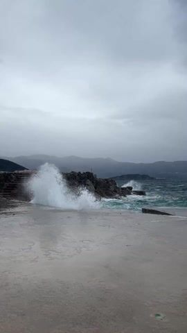The video shows a stormy seascape. A concrete pier or walkway is in the foreground, wet from the sea spray. To the left, dark, jagged rocks rise from the water, with waves crashing against them and sending white spray into the air. The water itself is a turbulent, dark teal color. In the background, a range of hazy mountains can be seen under a heavy, overcast sky. The overall atmosphere is bleak and stormy.