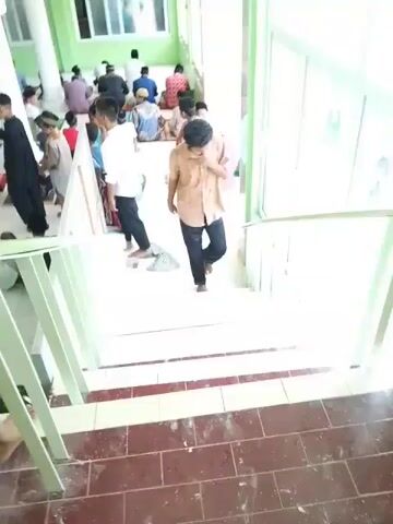 The video shows people inside a building. Many of them are seated on the floor of a room with white tile, presumably praying or in a gathering. Some people are walking up a set of stairs with white and red tiled treads and a green railing.  One person is descending the stairs, holding the railing. The room is brightly lit, with large windows allowing natural light to enter. The walls are painted green.