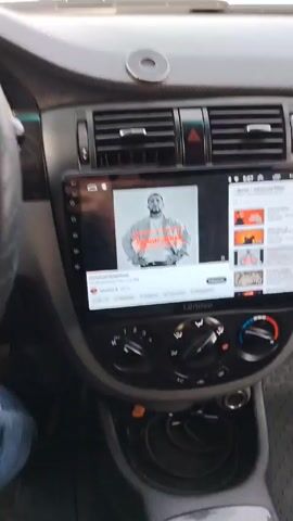 The video shows the interior of a car, particularly the dashboard and steering wheel. A Lenovo branded screen displays a music video on YouTube. The driver, presumably, is driving on a road with limited visibility. The speedometer shows the car is moving, but the exact speed is difficult to determine due to the angle and blur.