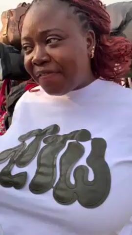 The video features a woman with braided, red-tinted hair, wearing a white t-shirt with a large olive-green logo on the front. She is looking to her right and speaking, with her hands moving as she gestures. Behind her is a display of bags and luggage. The setting appears to be outdoors in daylight.