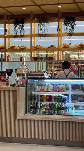 The video shows the interior of a cafe. A person in a white shirt and an apron is standing behind a counter, looking away from the camera. In front of the person is a coffee machine and a display case filled with drinks and some pastries. The shelves behind the counter are stocked with bread and other items, and there are hanging plants and lights. The counter itself is made of wood with vertical slats. The overall impression is a well-lit and modern cafe.