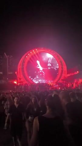 A concert crowd is visible in the foreground, facing a large circular stage with a dynamic light display. The stage features a prominent circular screen showing abstract red and white visuals, with figures of performers visible. Red stage lighting illuminates the scene, casting a vibrant glow over the audience and the stage setup. The overall atmosphere suggests an energetic live music performance at night.