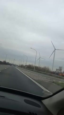 The video shows a view from inside a car driving on a highway. The road has multiple lanes and the weather is overcast.  Wind turbines are visible on the side of the road, interspersed with streetlights.  There are a few other cars on the road ahead.