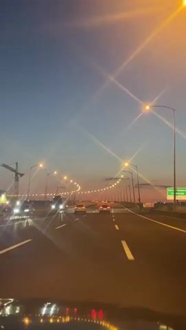 The video shows a car driving on a multi-lane highway at dusk. Streetlights illuminate the road, and other cars are visible in the distance. A crane can be seen to the left of the road. The sky is a mix of blue and orange.