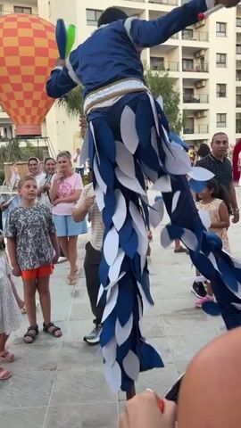 The video shows a street performer on stilts entertaining a crowd in an outdoor setting. The performer is dressed in a blue and white costume that resembles feathers, with a blue jacket and white and blue pants. He is juggling colorful objects, including red, green, and yellow clubs.

The crowd consists of people of all ages, including children who are watching with great interest. Some adults are smiling and appear to be enjoying the performance. The setting appears to be a resort or tourist area, with a large building in the background that has multiple floors and balconies. There is also a large hot air balloon in the background, which suggests a festive atmosphere. The time of day appears to be late afternoon or early evening, as the lighting is warm. The weather seems pleasant.

Notable details include the performer's height due to the stilts, the colorful juggling props, and the attentive audience. The overall mood is joyful and celebratory. There is no visible text in the video.