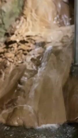 A video clip shows a close-up of a waterfall cascading down a rocky, muddy incline. The water is murky brown and appears to be flowing rapidly. The rocks are uneven and textured, with some areas looking smooth and others rough. The overall impression is one of a natural, perhaps underground, environment. There are no people visible in the clip. The lighting is dim, suggesting an indoor or cave-like setting.