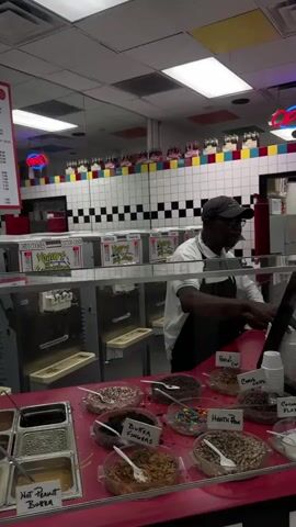 A man in a dark baseball cap and black apron stands behind a counter, preparing a dessert at a shop. The counter displays a variety of toppings, including hot peanut butter, butterfingers, M&Ms, and Heath bar pieces, in clear dishes with plastic spoons. Behind the counter are ice cream machines, and above them, a decorative border of white and black tiles, mirrored sections, and novelty bottle decorations. A menu board is visible to the left, and an "OPEN" sign hangs near the ceiling. The store's interior is brightly lit with fluorescent lights.